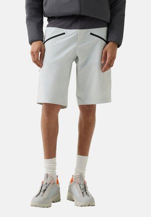 Man wearing light gray shorts with black zip pockets, white socks, gray and orange athletic shoes, and a dark jacket.