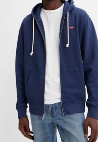 Marineblaue Zip-Hoodie mit Kapuze mit Kordelzug, weißen Akzenten, Fronttaschen und einem kleinen roten Logo auf der linken Brust. Über einem weißen T-Shirt getragen.