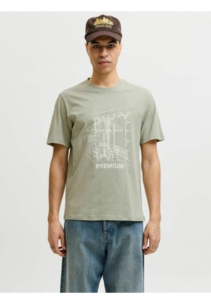 Mężczyzna w jasnooliwkowej koszulce z białym szkicem patio kawiarni i napisem „PREMIUM”, niebieskich dżinsach oraz ciemnobrązowej czapce z żółtym wzorem.