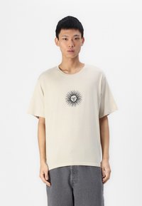 T-shirt beige a maniche corte realizzato in tessuto morbido, con un motivo di sole nero al centro. Collo a giroclassico, vestibilità rilassata, contraddistinta da maniche sezionate.