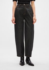 Jeans pretos de perna larga com um acabamento texturizado, apresentando costuras laranjas, fecho com botão e bolsos frontais. Combinados com botas pretas de tornozelo.