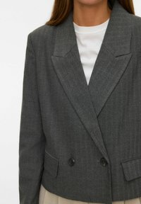 Sivi blejzer s uzorkom riblje kosti, dvostrukim kopčanjem, istaknutim reverima i dva dugmeta, uparen s bijelom košuljom ispod.