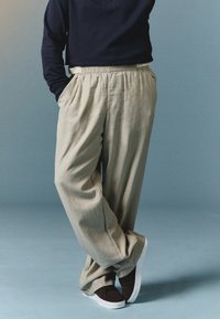 Pantaloni bej cu picioare largi, cu un finisaj texturat, având un brâu elastic și buzunare laterale, asortați cu pantofi slip-on de culoare maro închis.