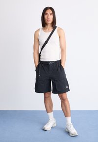 Man with long hair wearing white tank top, black cargo shorts with belt, white socks, sneakers, and a black crossbody bag standing on blue floor.