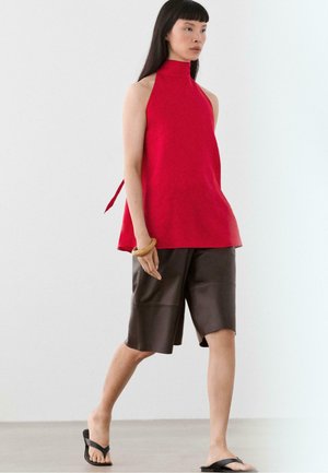 Frau trägt ein rotes ärmelloses Neckholder-Top, dunkelbraune knieumspielende Shorts, ein Holzarmband und schwarze Sandalen vor weißem Hintergrund.