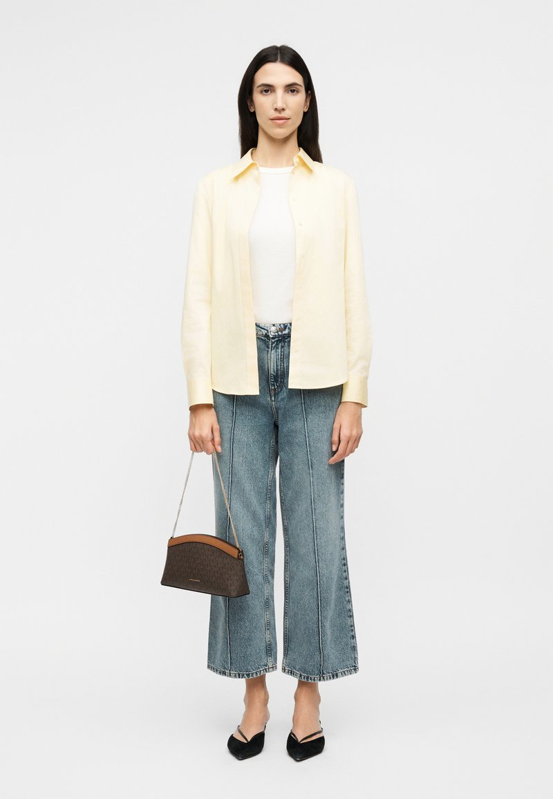 Γυναίκα με μακριά σκουρόχρωμα μαλλιά, φοράει ανοιχτό κίτρινο πουκάμισο με κουμπιά, λευκή μπλούζα, τζιν παντελόνι με φαρδιές κοντές πιέτες, μαύρα μαύρα παπούτσια με μυτερή μύτη, κρατώντας μια μικρή καφέ τσάντα.