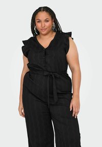 Crni kombinezon s V-izrezom, kratkim nabranim rukavima i vezanjem u struku. Sadrži teksturiranu tkaninu s suptilnim uzorkom i kopčanje na dugme.