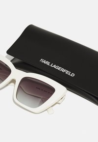 Bijele oversize sunčane naočale s gradijentnim tamnim sočivima, u pratnji crne zaštitne futrole s logotipom "Karl Lagerfeld" u srebrnoj boji.
