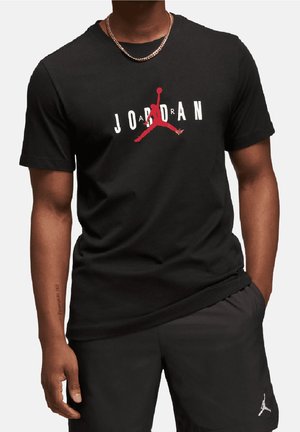 Czarny t-shirt Jordan z czerwonym logo Jumpman i białym tekstem noszony przez osobę o ciemnej karnacji, w połączeniu z czarnymi szortami z logo Jumpman.