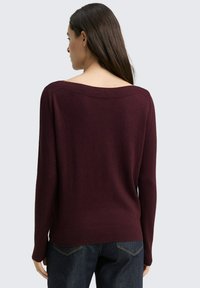 Vrouw met lang donker haar, gekleed in een donkerpaarse off-shoulder trui en donkerblauwe jeans, gezien vanaf de achterkant tegen een effen achtergrond.
