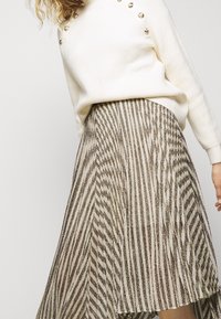 Žena v bílém svetru se zlatými knoflíky a metalickým pruhovaným skládacím sukní, pózující na jednobarevném světlém pozadí.