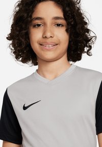 Maglia sportiva a maniche corte grigia e nera con tessuto testurizzato e design a V. Presenta un logo Nike nero sul petto.
