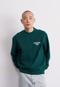 Mørkegrøn sweatshirt i bomuld med rund hals, ribbede ærmer og et lille hvidt logo på venstre bryst. Kombineret med lysegrå bukser.