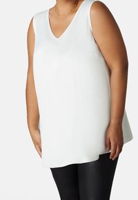 Vit ärmlös topp med v-ringad design, tillverkad av slätt tyg. Toppen har en avslappnad passform och når till mitten av höften.