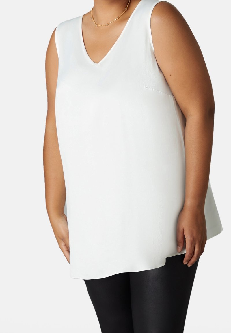 Vit ärmlös topp med v-ringad design, tillverkad av slätt tyg. Toppen har en avslappnad passform och når till mitten av höften.