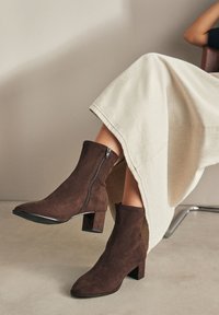 Hnedé členkové topánky zo semišu s špicatou špičkou, bočným zipsom a hrubým podpätkom. Nosené s krémovou sukňou, ktorá zdôrazňuje textúru a dizajn.