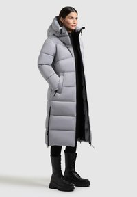 Cappotto a vento lungo di un grigio chiaro, con cappuccio, dotato di chiusura con zip, tasche laterali e una texture trapuntata. Abbinato a stivali neri con lacci.