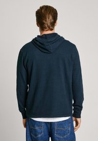 Kapucēts džemperis tumši jūras zilā krāsā, izgatavots no mīksta kokvilnas. Ar kenguru kabatu un ribotām piedurknēm, valkāts virs ziliem denim džinsiem.