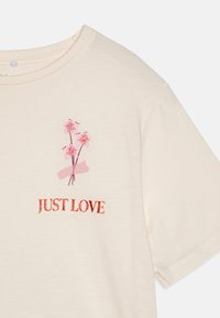 T-shirt crème clair avec un col rond. Doté d'un design floral rose et du texte "JUST LOVE" en rouge brillant. Matière en coton doux.
