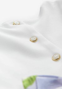 Tessuto bianco con texture a coste, caratterizzato da bottoni tono oro con centri simili a perle. Include accenti colorati in verde e blu.