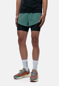 Grüne Sportshorts über schwarzen Kompressionsshorts, kombiniert mit mehrfarbigen Sneakers und weißen Socken mit Muster.