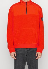 Punainen pusero, jossa on korkea kaulus, puolivetoketju edessä ja teksturoitu fleece-vartalo. Ominaisuuksiin kuuluu resorireunus helmassa ja hihansuissa sekä etutasku.