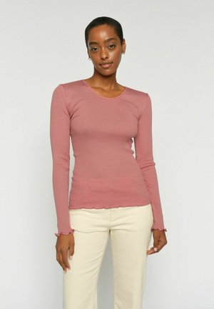 Žena s krátkymi vlasmi, ktorá má na sebe priliehavé ružovkasté tričko s dlhým rukávom a svetlo béžové nohavice, stojaca pred jednoduchým svetlým pozadím.