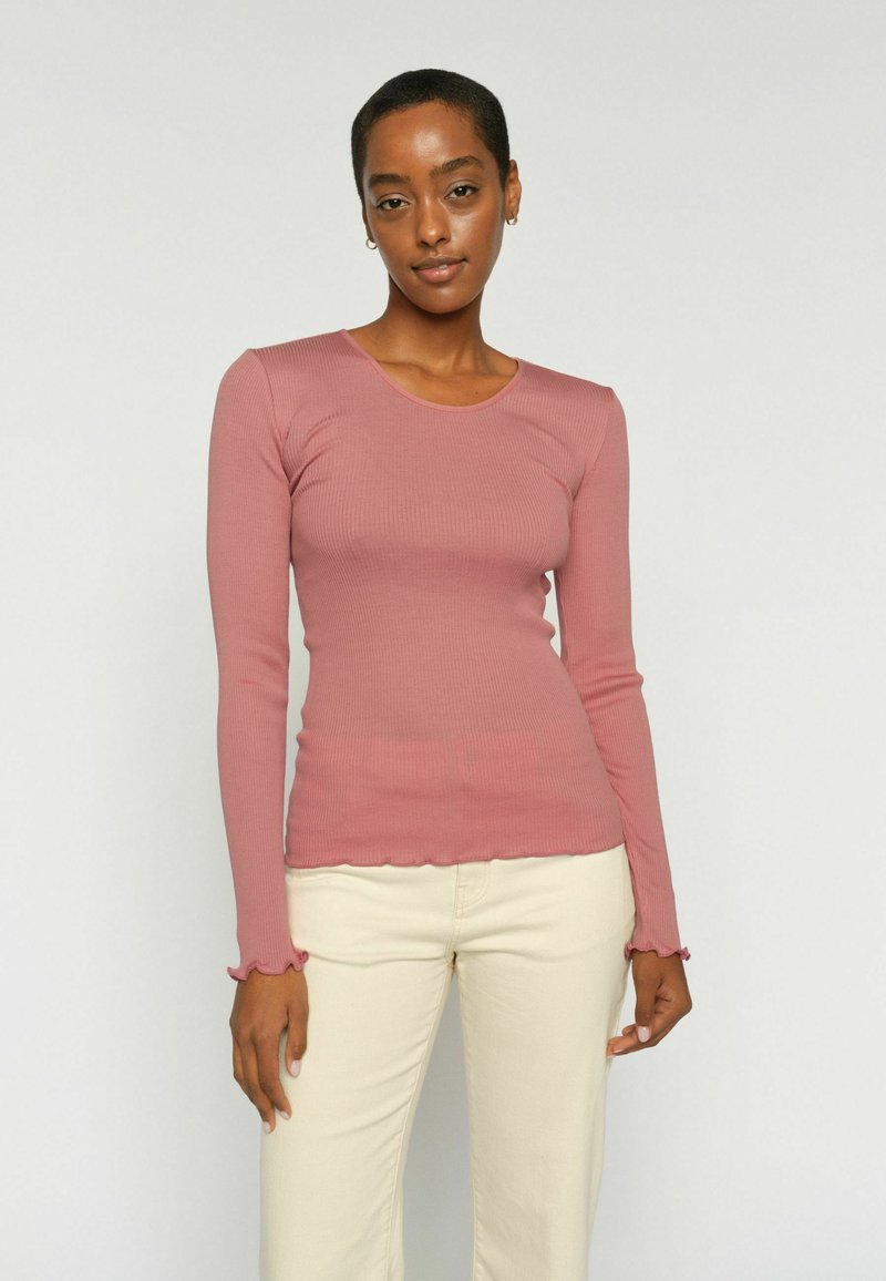 Femme aux cheveux courts portant un haut ajusté à manches longues rose poudré et un pantalon beige clair, debout devant un fond clair uni.