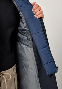 Granatowa kurtka z pikowanym wnętrzem, z ciemną wzorzystą podszewką, kieszenią na zamek oraz teksturowanym zewnętrznym materiałem, pokazująca dłoń chwytającą suwak.