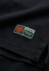 Donker stof met een groen en rood rechthoekig label genaamd "Superdry Vintage Athletic Outfitters" dat nabij een zoom is genaaid.