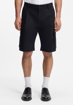 Bărbat purtând pantaloni scurți cargo negri, șosete albe până la mijlocul gambei și pantofi eleganți negri, stând în picioare pe un fundal alb simplu.