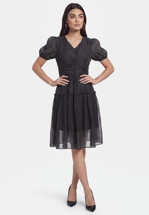 Frau trägt ein knielanges schwarzes Kleid mit Puffärmeln, Knopfdetails und transparentem Stoff, steht mit den Händen in der Hüfte und schwarzen Absätzen.