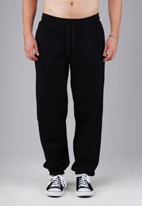 Man wearing black sweatpants with drawstring, black and white sneakers, standing against a plain gray background.