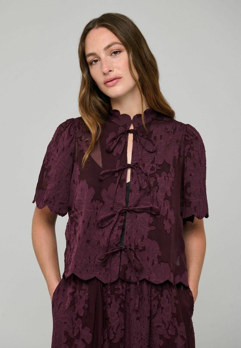 Femme aux longs cheveux ondulés porte un chemisier en dentelle florale transparente violet foncé avec des manches courtes et des nœuds sur le devant.