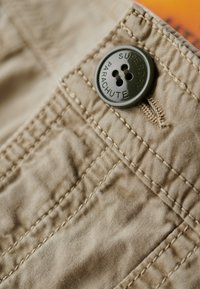 Ciemnozielony guzik przyszyty do beżowej tkaniny z widocznym przeszyciem, guzik grawerowany napisem "SUPERDRY PARACHUTE".