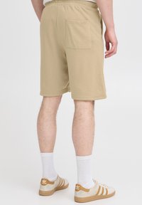 Beige cotton shorts with an elastic waistband and a back pocket. Model wears white socks and beige sneakers with white stripes.