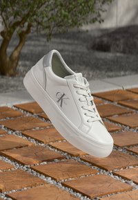 Witte leren sneakers met een glanzende afwerking, vetersluiting, grijze accenten en een logo aan de zijkant, gepresenteerd op een gestructureerd houten oppervlak.