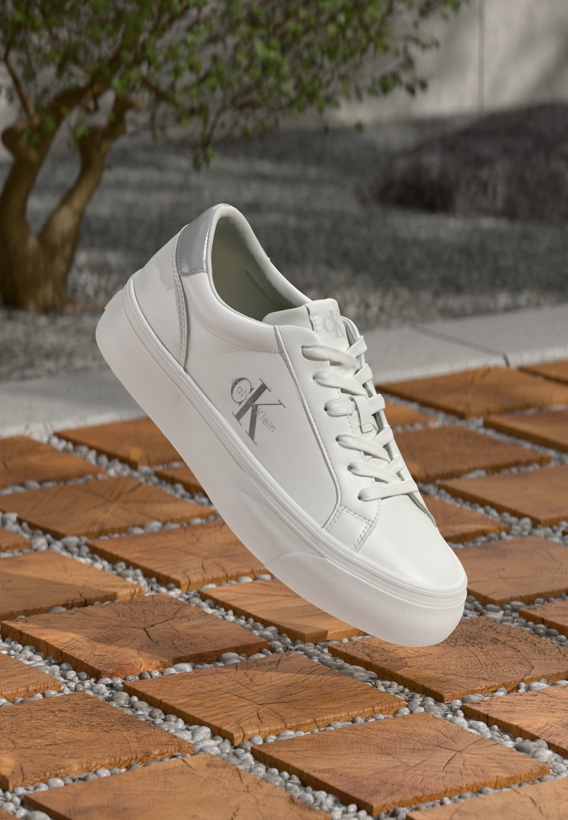 Witte leren sneakers met een glanzende afwerking, vetersluiting, grijze accenten en een logo aan de zijkant, gepresenteerd op een gestructureerd houten oppervlak.