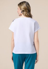Witte shirt met korte mouwen en een relaxte pasvorm, met een decoratieve accent op de schouder en een gladde, zachte textuur. Gecombineerd met teal gekleurde broek.