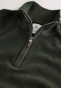 Pull en tricot vert foncé avec une demi-fermeture éclair et un col haut côtelé, orné d'une petite languette en tissu sur la fermeture éclair.