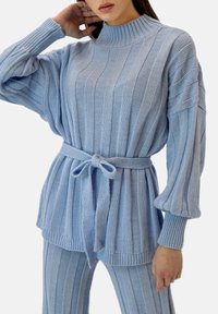 Jasnoniebieski sweter z dzianiny o prążkowanej fakturze, z wysokim kołnierzem i szerokimi rękawami; posiada pasek w talii umożliwiający regulację dopasowania.