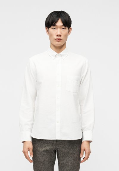 Jeune homme portant une chemise blanche à boutons avec une poche poitrine et un pantalon gris, debout devant un fond clair uni.