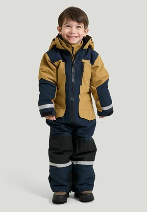 Smilende ung dreng iført en brun og navy vinterjakke og bukser med reflekstriber, stående foran en ensfarvet baggrund.