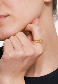 Goldene Doppelring mit einem gedrehten Knotendesign, das eine glatte Textur und ein glänzendes Finish aufweist. Von Hand präsentiert, sanft gegen die Gesichtshaut gelehnt.