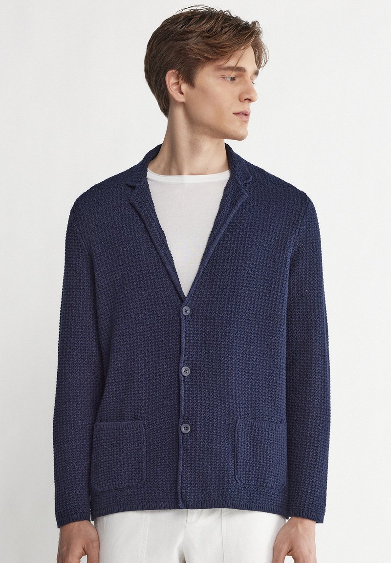 Jeune homme portant un cardigan bleu marine texturé boutonné par-dessus un t-shirt blanc à col rond, regardant vers sa droite.
