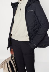 Giacca a vento nera con cappuccio, dotata di chiusura con zip e motivo trapuntato. Indossata sopra un maglione in pile chiaro e pantaloni scuri.