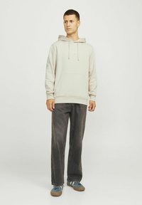 Sudadera beige claro con bolsillo frontal y cordones, combinada con pantalones de pierna ancha gris oscuro y zapatillas azules. Diseño simple y corte relajado.