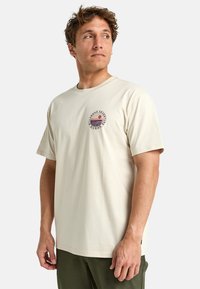 Camiseta crema de algodón, con un gráfico circular que presenta un diseño de atardecer y texto. Mangas cortas y corte relajado.