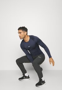 Marineblaues Langarm-Sportshirt mit Rundhalsausschnitt, kombiniert mit grauen, schmalen Hosen. Schwarze Sneaker mit weißen Akzenten runden das Outfit ab.