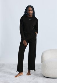 Črni komplet majice z dolgimi rokavi in trenirke z majhnim belim logotipom. Tkanina z teksturo, sproščen kroj, z nabranimi spodnjimi robovi hlač s povodcem.